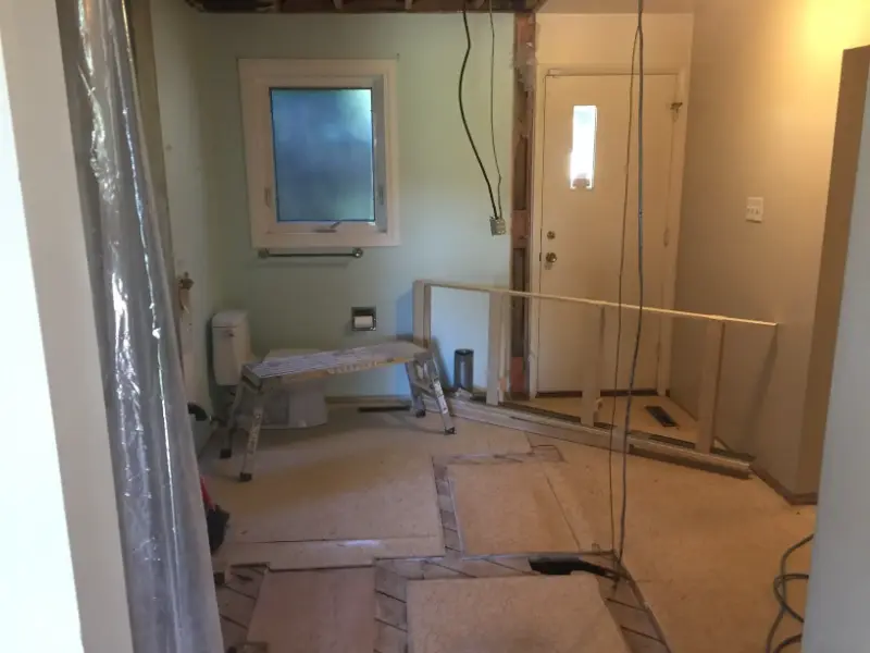 bathroom-renovation-in-progress A room under renovation with exposed wiring and walls. A step ladder is placed in the center, and the flooring is partially removed. A window and door are visible, and construction materials are scattered around.