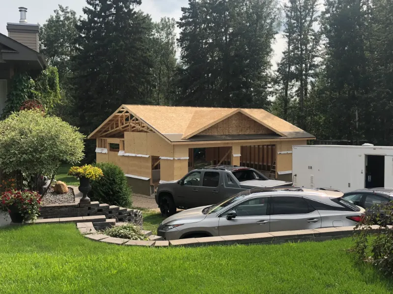 building-frame-with-vehicles-in-yard A partially constructed garage stands in a residential area, surrounded by lush greenery and trees. Two vehicles are parked in the driveway, and a trailer is situated beside the garage. A well-manicured lawn and garden are in the foreground.