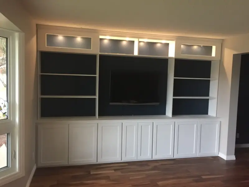 built-in-entertainment-unit-with-tv A built-in entertainment unit with white cabinets and open shelves surrounds a mounted TV on a dark background. Recessed lighting highlights the upper shelves. The unit is set against a wood floor and adjacent to a large window.