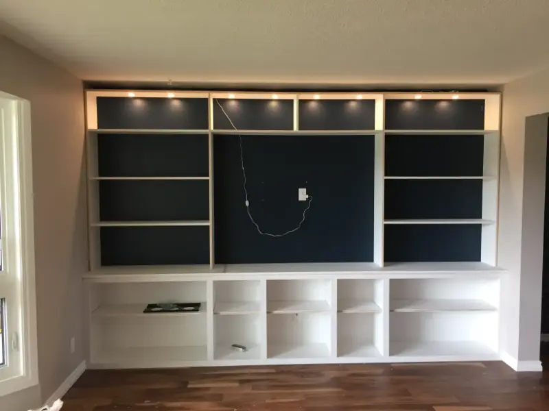built-in-shelves-with-lighting A wall-mounted white entertainment center with open shelving, dark blue paneling, and built-in lighting. There is an unconnected wire and an empty wall mount space for a TV. The unit stands on a hardwood floor.