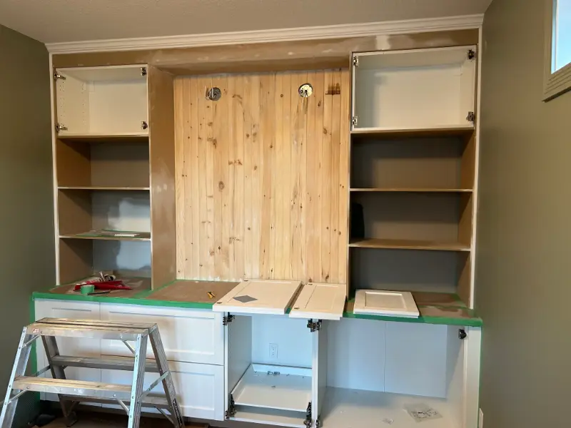 cabinetry-wall-construction A partially installed wall unit with wooden backing and white cabinets. The unit features open shelves and empty spaces for cabinet doors. A small ladder and tools are in front of the unit, and a protective cover is on the lower countertop.