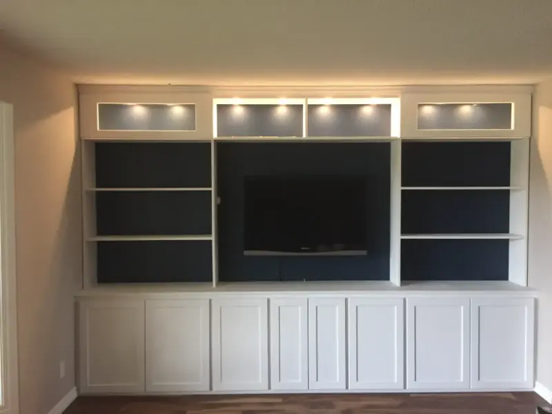 completed-entertainment-unit-with-tv A built-in entertainment center with white cabinetry, open shelves on either side, and a mounted flat-screen TV in the center. The upper shelves have backlighting, and the lower cabinets have closed doors.