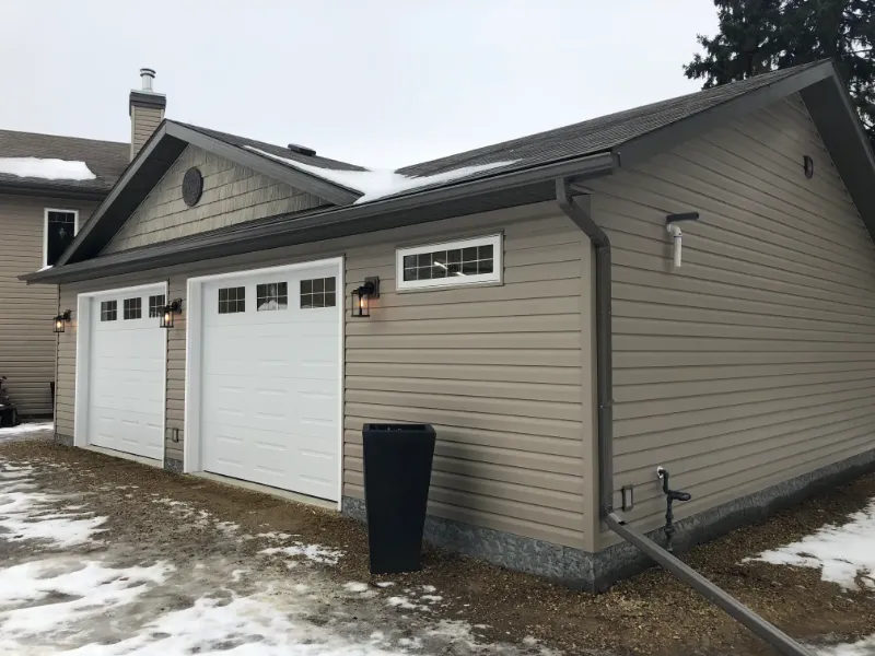 completed-garage-with-double-doors A beige detached garage with two white sectional doors, each featuring a small row of windows. The garages exterior includes lantern-style lights above each door. Sparse snow covers the ground and the roof, with pine trees visible in the background.