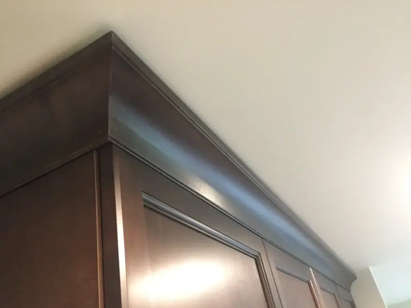 dark-cabinet-with-crown-molding-detail Close-up view of the top edge of a kitchen cabinet, featuring dark wood with a smooth finish and a crown molding design just below a white ceiling. The cabinet door has a rectangular panel design. Natural light illuminates the area slightly.