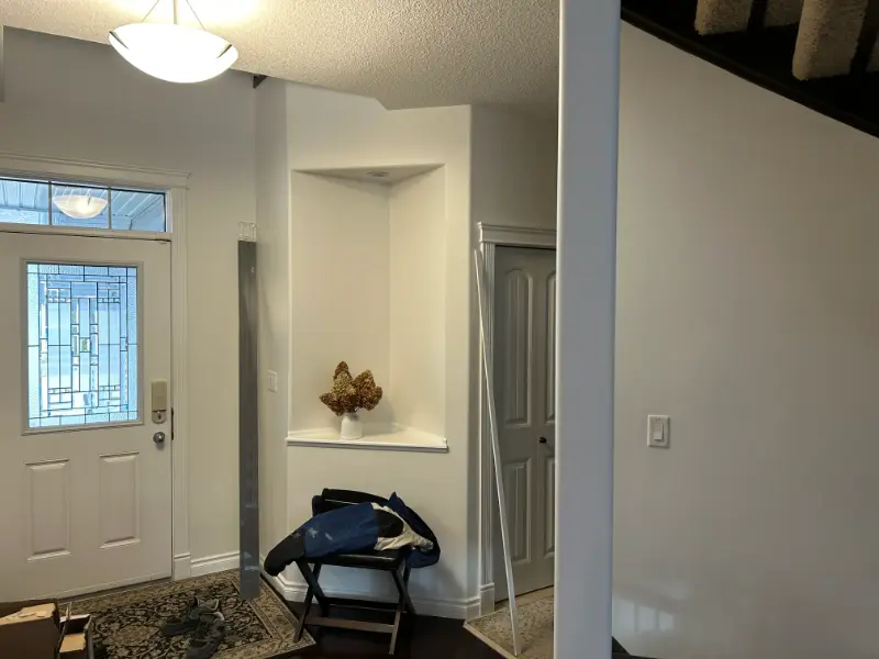 entryway-corner-display-niche A hallway featuring a decorative stained glass door, a small nook with a vase of dried flowers, a folded chair, and a gray coat. The floor is dark wood with a patterned rug, and a staircase ascends to the upper level.