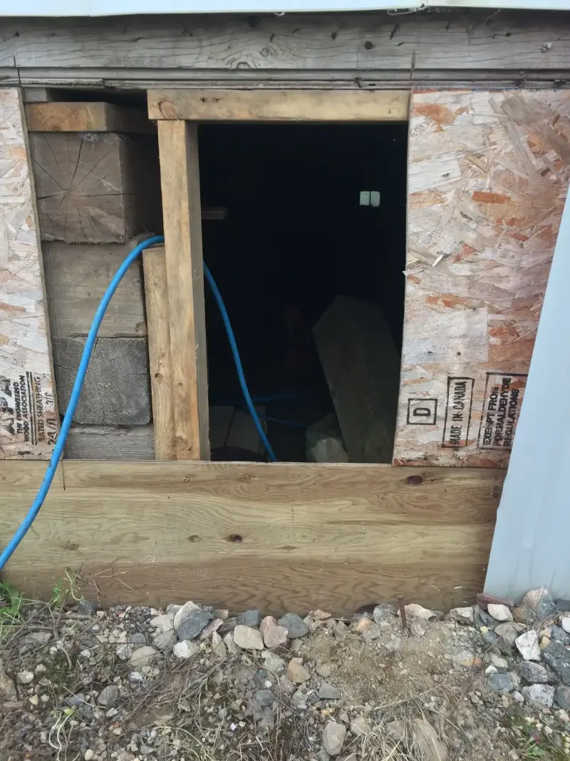 exterior-basement-access-construction A small rectangular opening in a wooden structure, surrounded by a mix of plywood and wood planks. A blue hose passes through the opening. The ground is covered with gravel and small stones.