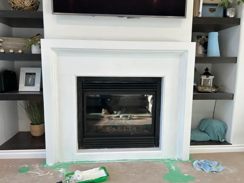 fireplace-mantel-painting A white fireplace with a black interior, surrounded by built-in shelves. The shelves hold decorative items like plants, a lantern, and framed photos. The floor is covered with a tarp and painting supplies, with green paint on the floor.