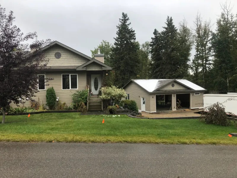 front-view-house-and-garage-addition A single-story house with beige siding and a matching detached garage. The front yard features green grass, bushes, and small trees. A paved driveway leads to the garage, and orange markers are placed on the lawn. Tall trees are in the background.