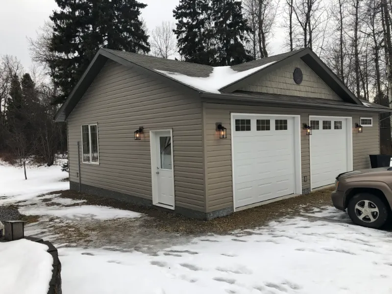 garage-exterior-with-side-door-and-snow A beige house with a snowy roof and ground, featuring two white garage doors and a side door. Outdoor wall lights are on, surrounded by leafless trees and a parked car partially visible on the right.