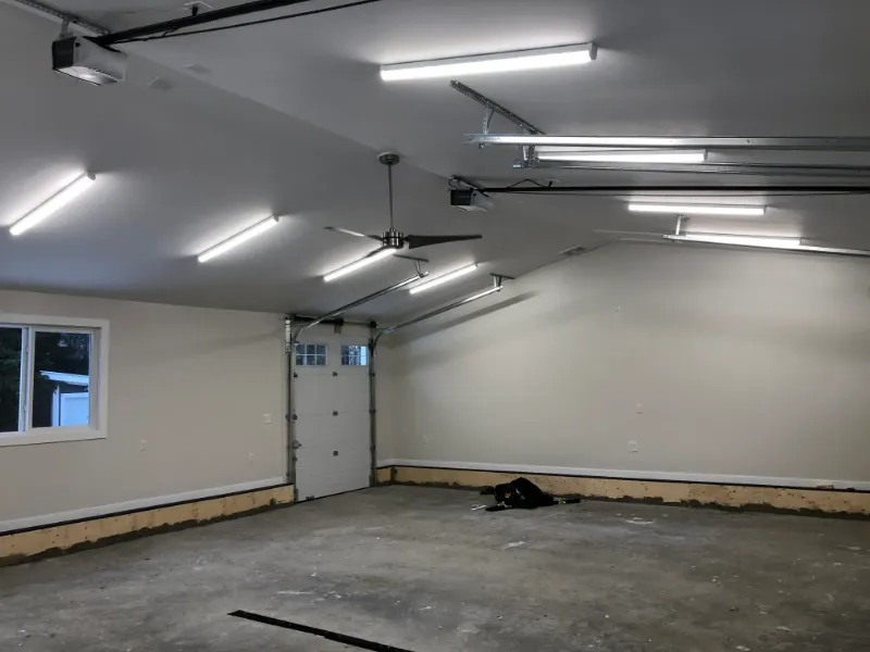 garage-interior-with-lighting-and-ceiling-fan A spacious, empty garage with concrete floors and off-white walls. The ceiling features fluorescent lights and a ceiling fan. A roll-up door with windows is visible on the left, and some tools are scattered on the floor.