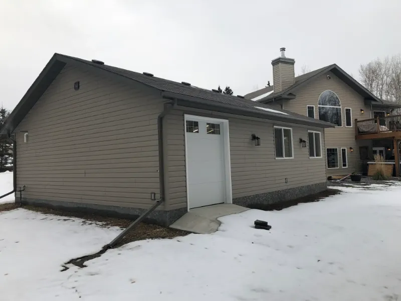 house-addition-during-winter A beige house with a two-story section and a single-story extension. The yard is partially covered in snow, and bare trees are visible in the background under a cloudy sky. A wooden deck is seen on the right.