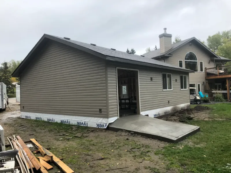 house-addition-with-concrete-ramp A newly constructed beige garage with a sloped roof and partially open door is next to a two-story house. The ground is bare with some building materials scattered around under an overcast sky.