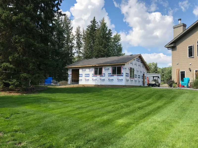 house-extension-with-tyvek-wrap A small house under construction is wrapped in Tyvek, surrounded by trees. Its next to a finished house with beige siding. In front, there’s a lush green lawn under a blue sky with clouds. A few outdoor chairs are nearby.