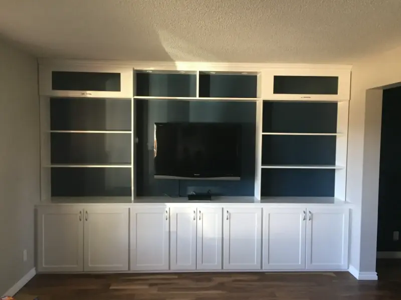 illuminated-built-in-unit-with-tv A built-in white entertainment center with a mounted flat-screen TV in the middle. The unit features open shelves on either side of the TV and closed cabinets below. The floor is hardwood, and the walls are light-colored.