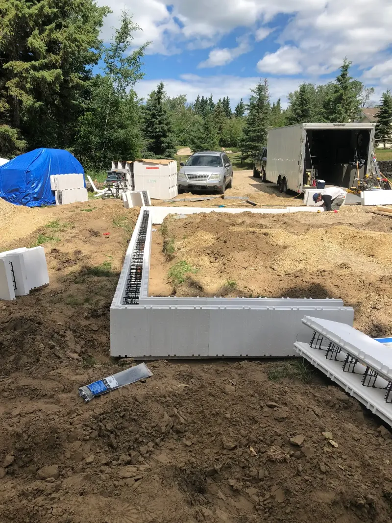 insulated-foundation-framing-in-yard Construction site of a building foundation with white insulated concrete forms, surrounded by soil. A truck and trailer are in the background, next to trees and a blue tarp covering materials under a blue sky.