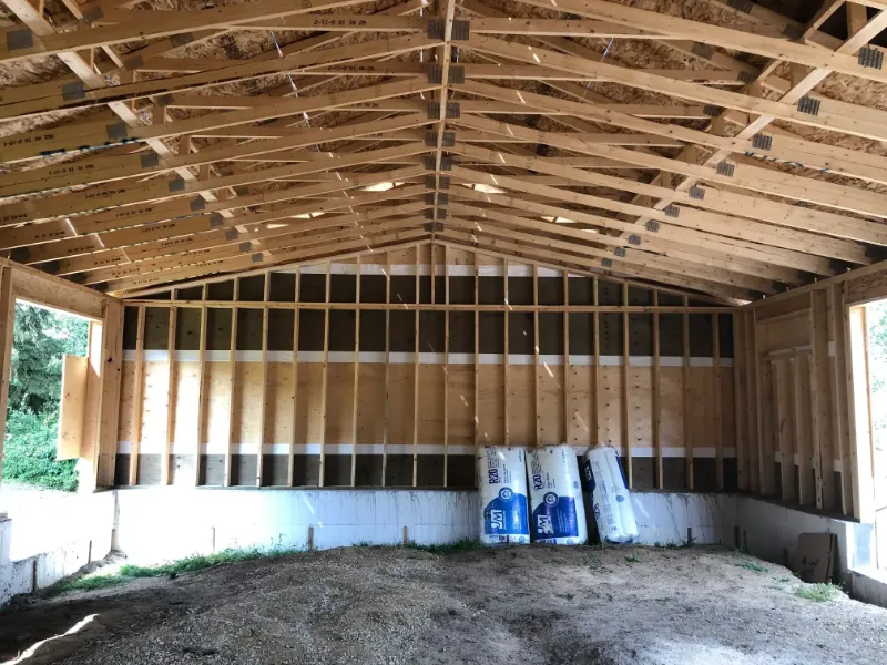 interior-framing-with-insulation-supplies Interior view of an unfinished wooden structure, featuring exposed beams and walls with partial insulation. Several rolls of insulation are stacked on the right. The floor is gravel, and natural light streams in from the open side walls.