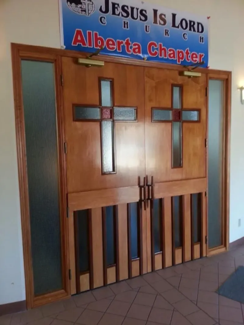 jesus-is-lord-alberta-wooden-doors Wooden church doors with cross-shaped glass panels under a "Jesus Is Lord Church Alberta Chapter" sign.