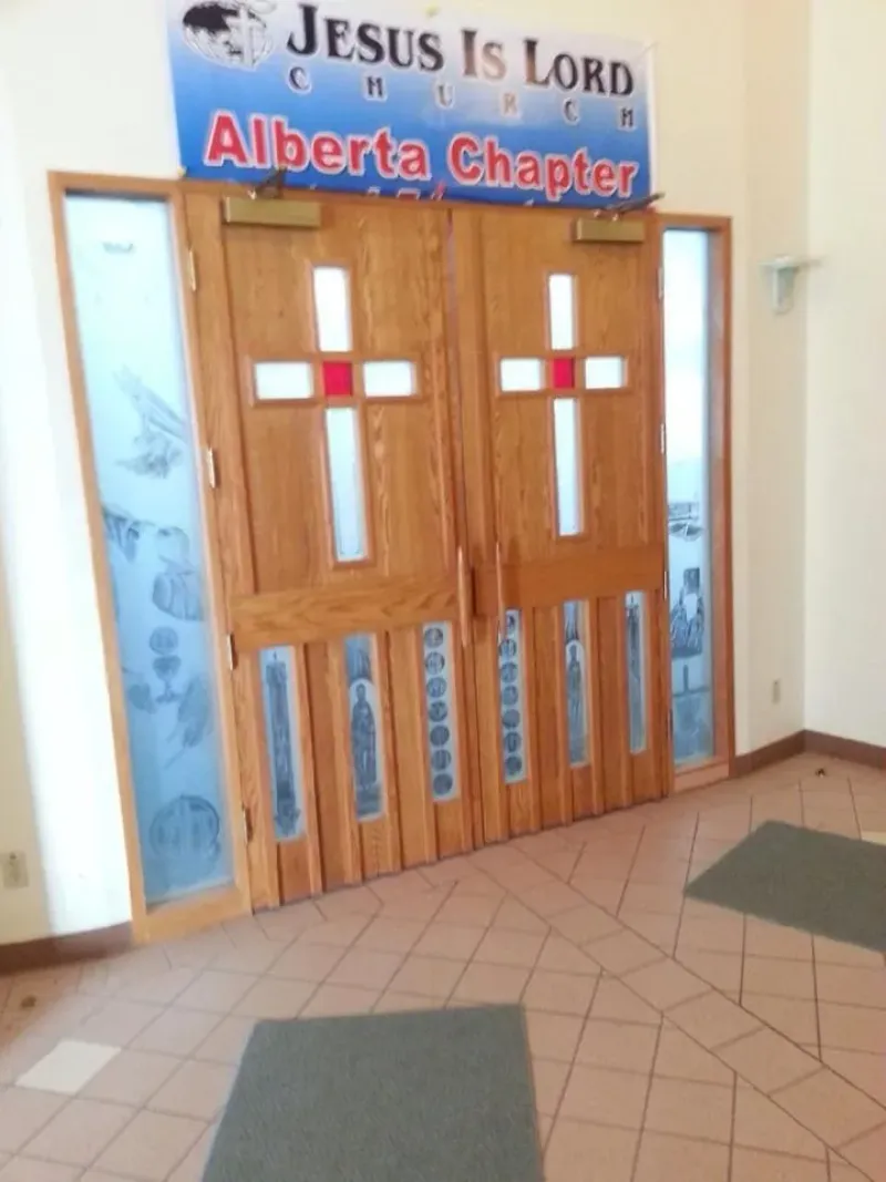 jesus-is-lord-church-alberta-decorative-doors Wooden church doors with decorative glass panels and cross-shaped designs beneath a "Jesus Is Lord Church Alberta Chapter" sign.