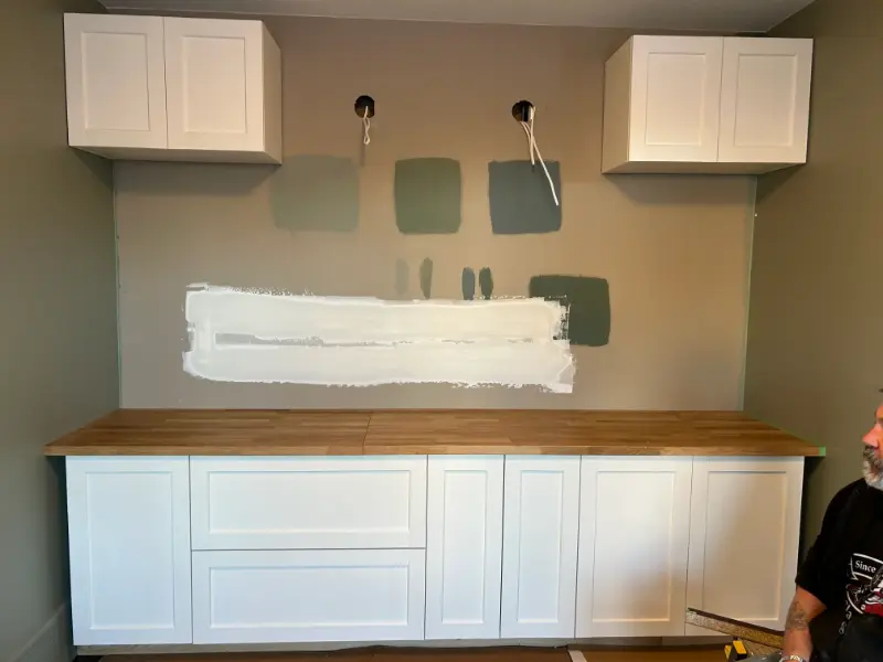 kitchen-paint-samples-cabinets A kitchen wall with several paint swatches in various shades of green and gray above a wooden countertop. White cabinets are installed above and below the counter. There is a person seated in the lower right corner.