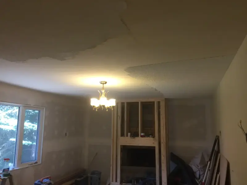 living-room-ceiling-repair-and-fireplace-framing A dimly lit room with a chandelier, unfinished walls, and a partially constructed fireplace. The ceiling shows signs of water damage with peeling paint. A window on the left lets in some natural light. Construction materials are scattered around.