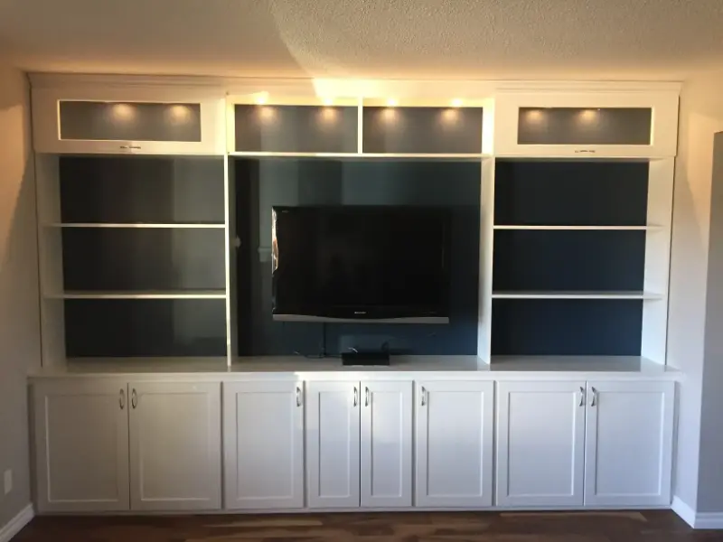 modern-built-in-shelving-unit Built-in white entertainment center with shelves and cabinets, positioned against a wall. A flat-screen TV is centrally located, surrounded by open shelving. Upper sections feature dim lighting, adding a subtle glow to the unit. Hardwood flooring below.