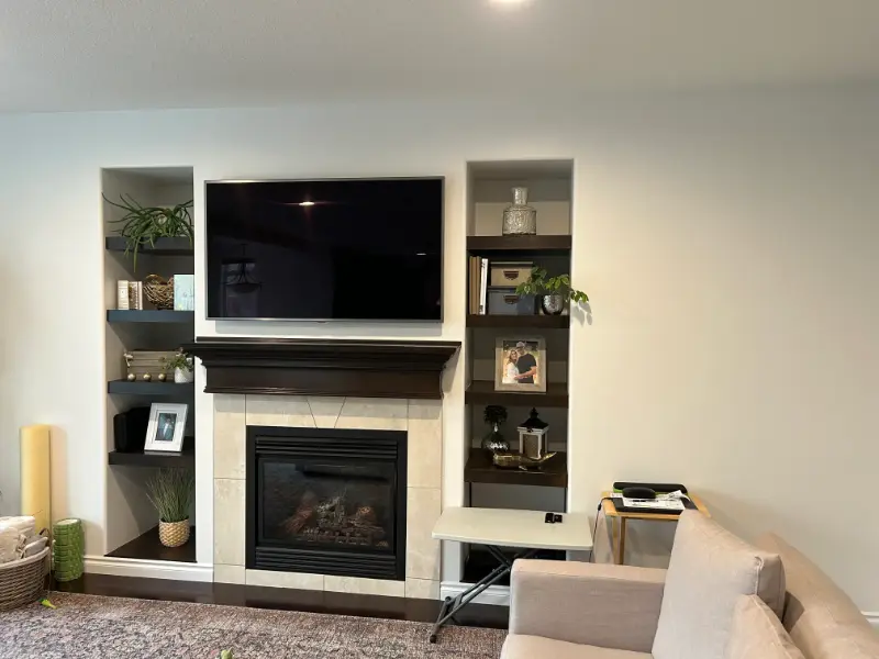 modern-fireplace-with-shelving A living room with a TV mounted above a fireplace, flanked by built-in shelves holding plants and decor. A beige couch is in the foreground, and a small desk with a chair is positioned next to the right shelf.