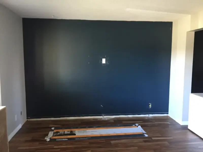 navy-accent-wall-preparation A room with a dark blue accent wall, featuring a light switch and power outlet. The floor is wooden, and a long piece of packaging rests on the ground. The room is empty, with white walls on the sides and a bright section on the right.