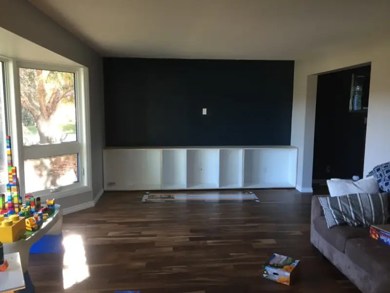 playroom-with-built-in-shelving-and-toy-table A room with a wooden floor, dark accent wall, and white built-in cabinets. A bay window with a view of trees is on the left. A beige sofa with a striped pillow is on the right. Toy blocks are scattered on the floor and table.