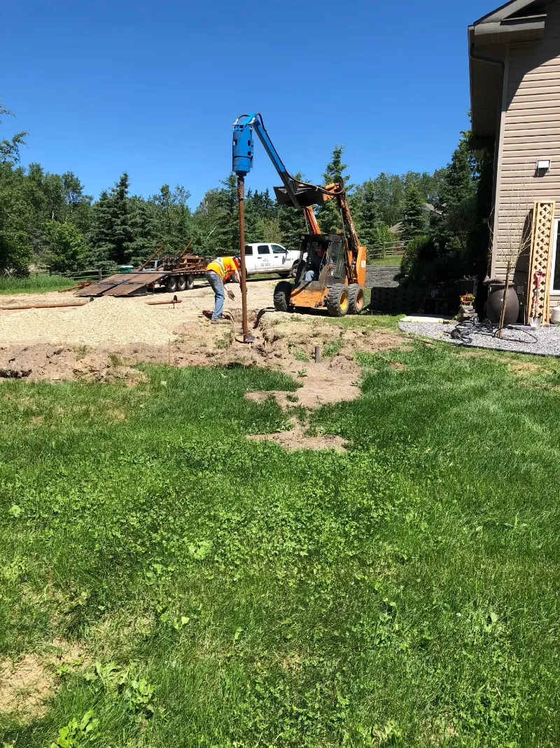 post-installation-with-mini-excavator A person in an orange safety vest operates a small construction vehicle with an auger attachment, drilling into the ground. Another worker stands nearby. The site is next to a house, with a grassy lawn and trees in the background.