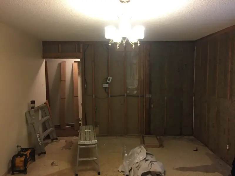 room-under-renovation-with-exposed-insulation A dimly lit room undergoing renovation, with stripped walls revealing wooden studs. A ladder and some tools are on the floor, while a chandelier hangs from the ceiling. Plastic sheets and materials are scattered around.