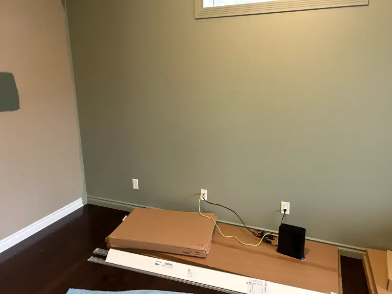 room-with-green-accent-wall A sparsely decorated room with beige walls and dark wooden floors. A modem/router is placed on a cardboard box on the floor, with cables connected to it. A small window is visible near the ceiling.