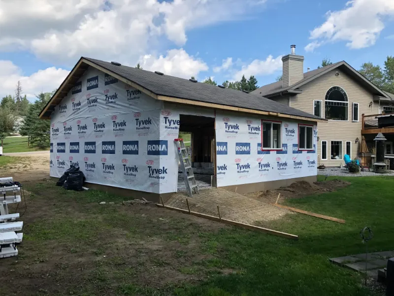 side-view-of-house-extension-with-tyvek A house under construction with Tyvek insulation wrap visible on the exterior. A ladder leans against the building, which is set in a grassy area with a clear sky and trees in the background. Construction materials are scattered nearby.