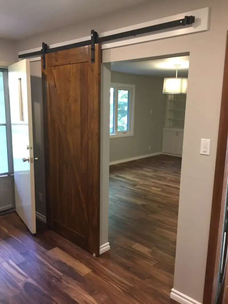 sliding-barn-door-installed-in-room A room with wooden flooring features a sliding barn door on a rail. The door is partially open, leading into an adjacent room with a large window and overhead light fixture. Walls are painted off-white.