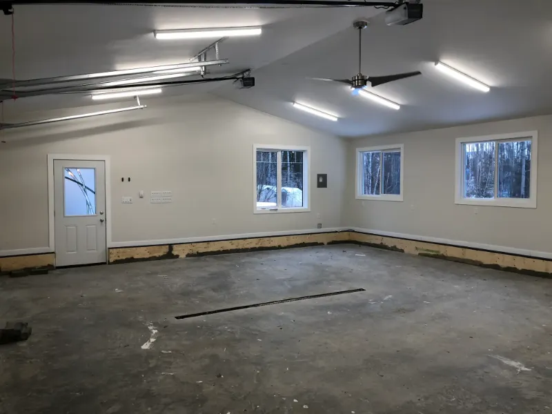 spacious-garage-interior-with-windows A spacious, empty garage interior with unfinished flooring, white walls, and bright overhead lighting. There are three windows, a ceiling fan, and a white door on the left. The room appears clean and ready for further development.