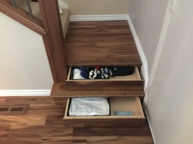 stairs-with-hidden-storage-drawers Two wooden staircase steps have built-in pull-out drawers. The top drawer is partially open, revealing folded clothing, while the bottom drawer is fully open, showing more items inside. The stairs lead up to a carpeted area.