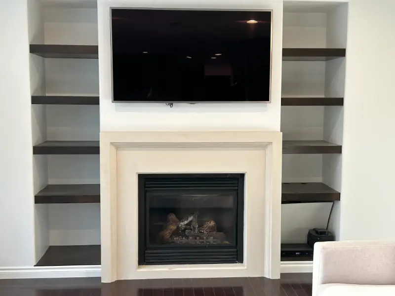 tv-fireplace-shelves A modern living room features a wall-mounted flat-screen TV above a sleek fireplace. The fireplace is set within a light-colored mantel. Built-in wooden shelves flank both sides, and a chair is partially visible in the foreground.