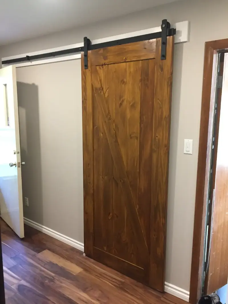 wooden-barn-door-closed-in-room A wooden sliding barn door with a dark metal rail is installed in a room with light-colored walls and wooden flooring. The door is partially open, revealing a glimpse of the adjacent space.
