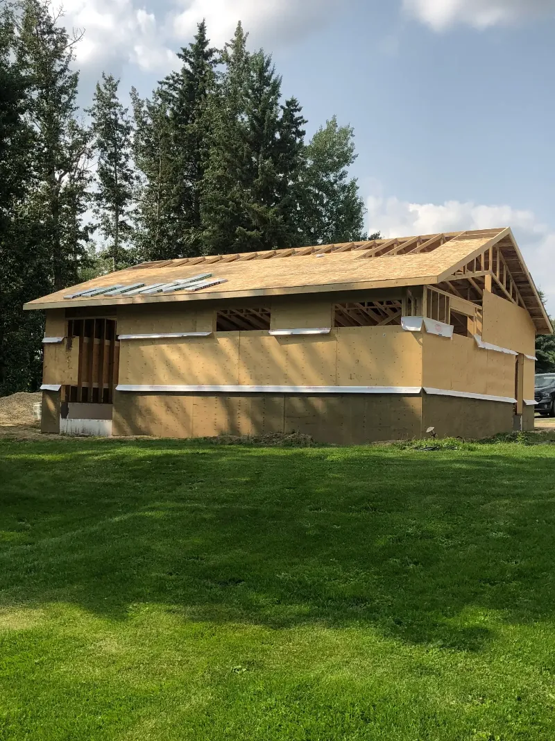 wooden-frame-building-with-roof-sheathing A partially constructed wooden house with exposed beams and a roof framework, set in a grassy area surrounded by tall trees under a partly cloudy sky.