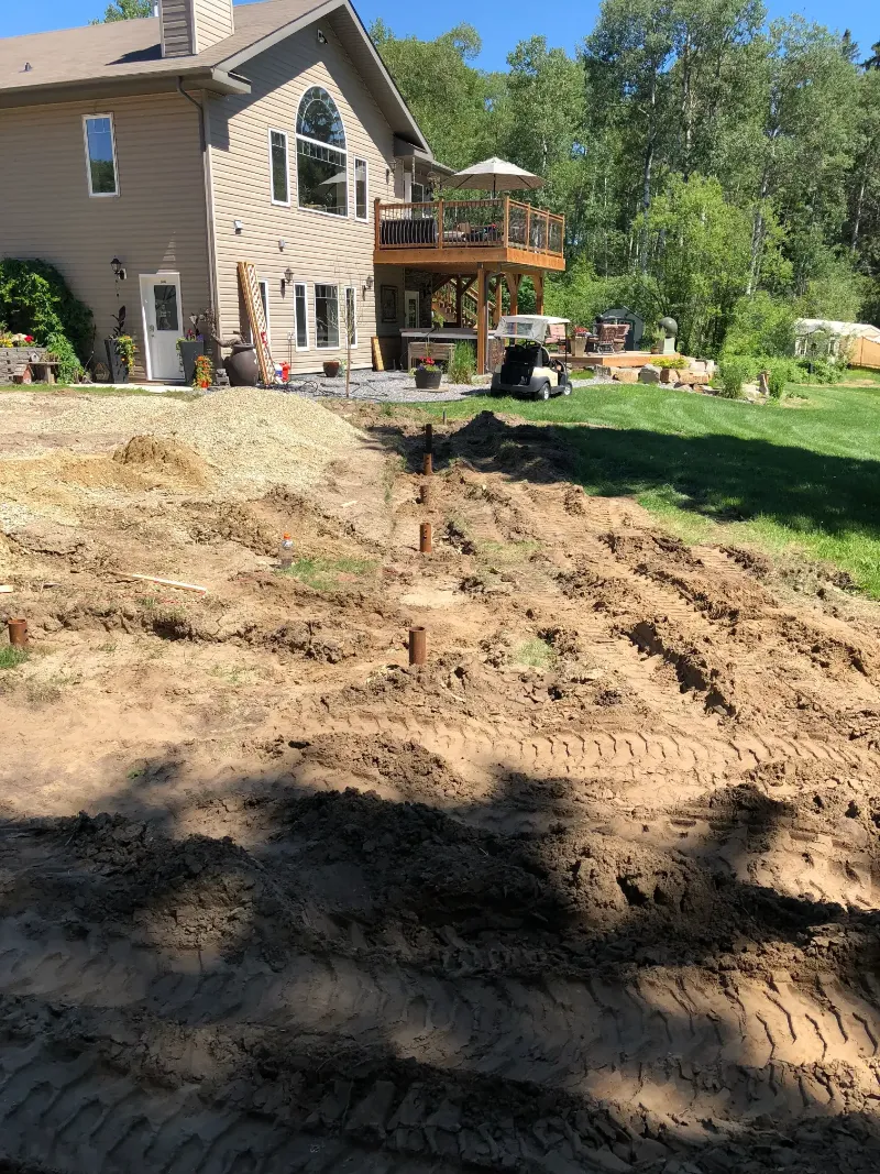 yard-with-trenches-and-installed-posts A backyard with freshly dug soil and visible tire tracks, leading toward a modern house. The house has large windows and a wooden balcony. There are trees and greenery surrounding the area.